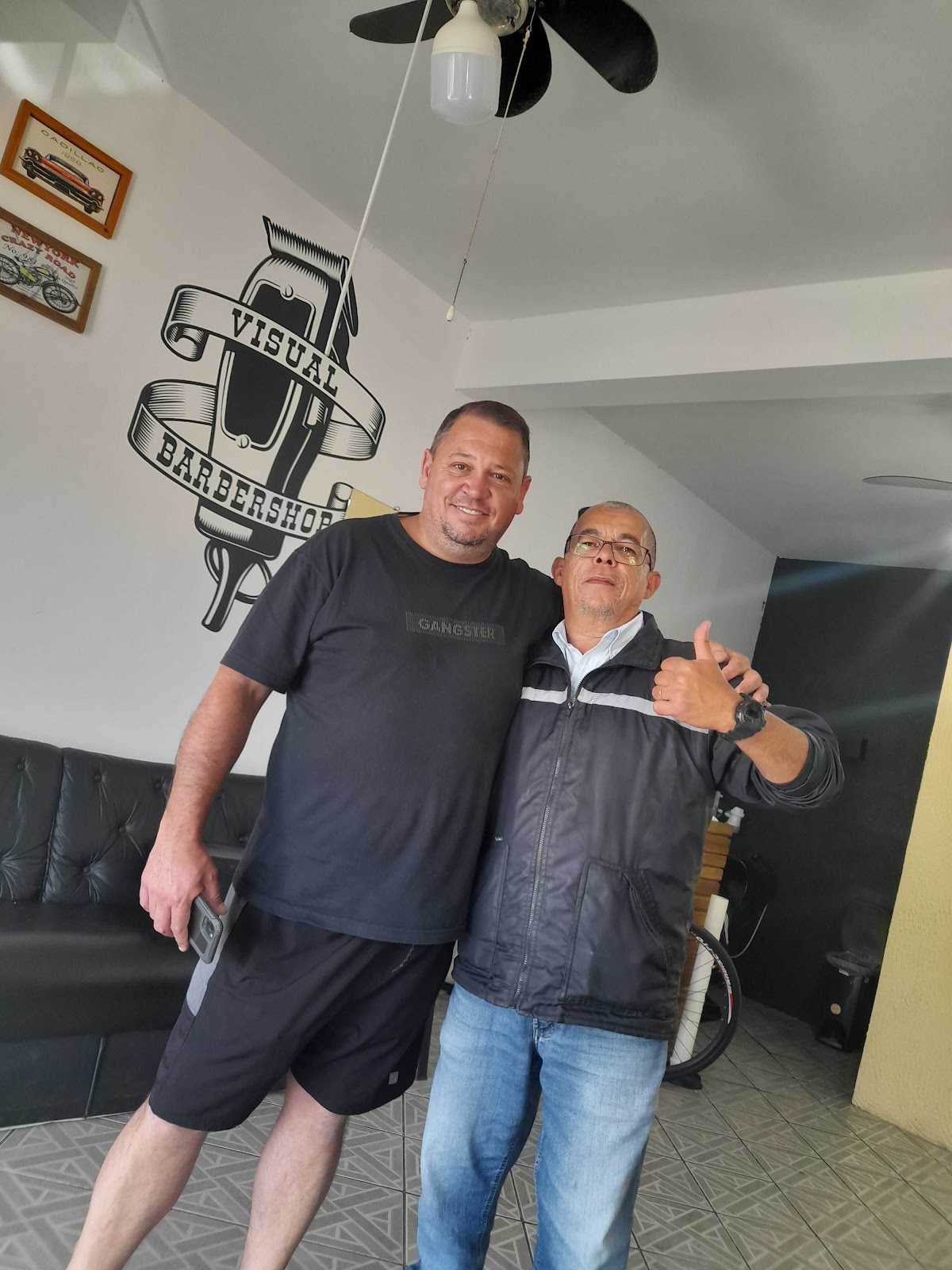 Cliente e barbeiro posando ao lado do logo Visual Barbershop pintado na parede